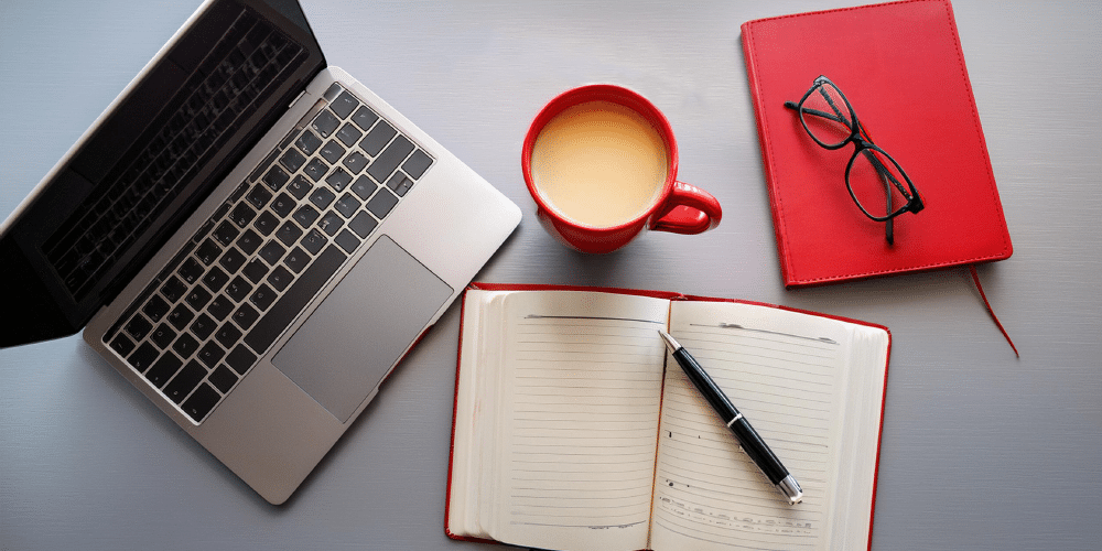 Looking down on an open laptop, cup of coffer, notebook, pen, and glasses.