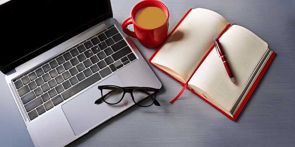 An open laptop has a cup of coffee and a notebook and pen beside it. A pair of glasses sits on the keyboard.